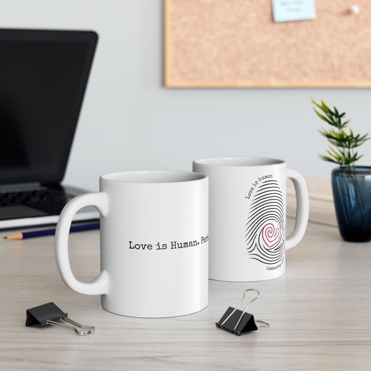 Two white ceramic mugs on a light wood desk in a styled office setting. The front mug reads 'Love is Human.