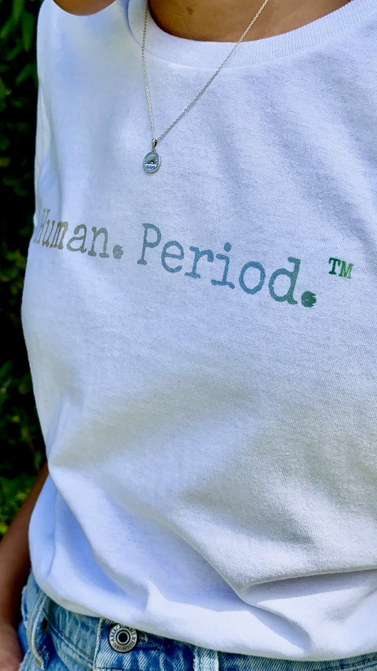A close-up of a person wearing a white crewneck t-shirt with the words 'Human Period.' printed across the chest in muted green and blue-gray tones, paired with blue jeans and a silver pendant necklace.