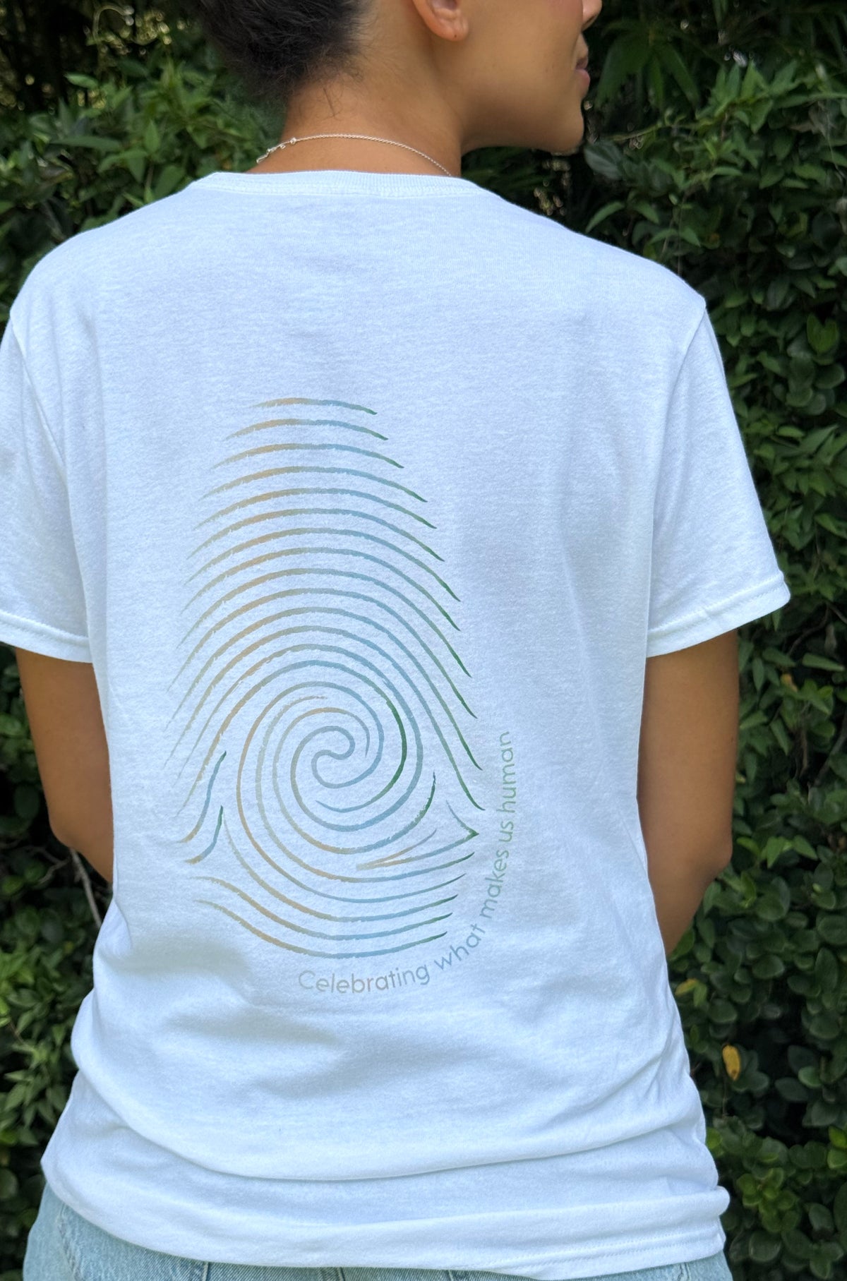 A person shown from behind wearing a white t-shirt with a large rainbow-colored spiral fingerprint graphic on the back, set against a background of green foliage.