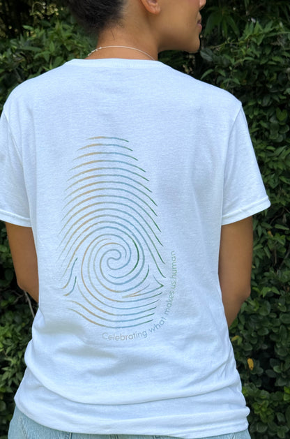 A person shown from behind wearing a white t-shirt with a large rainbow-colored spiral fingerprint graphic on the back, set against a background of green foliage.