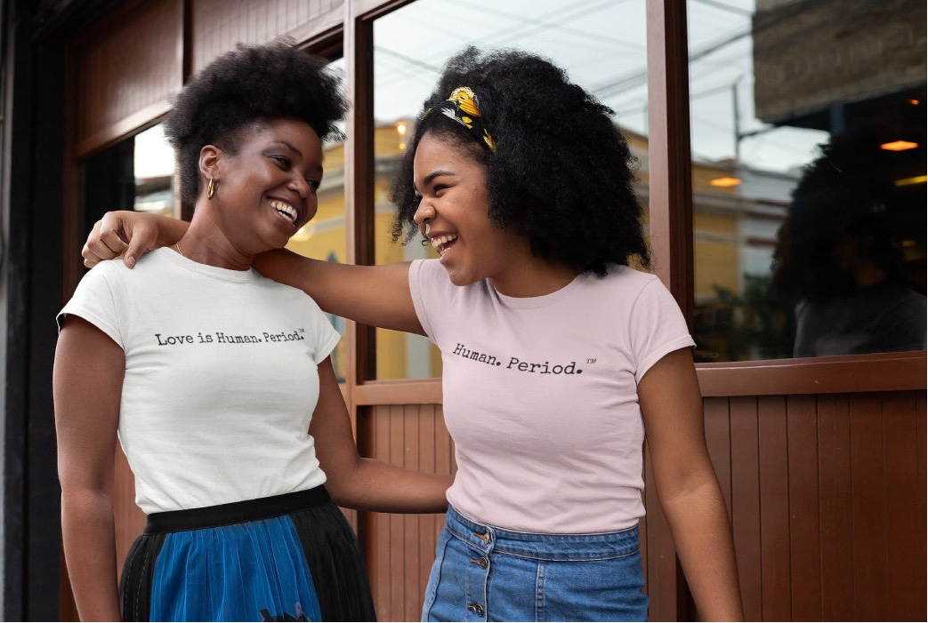 Two women laughing together in front of a storefront window. Both are wearing matching light-colored graphic T-shirts with the text 'Love in Human, Period.
