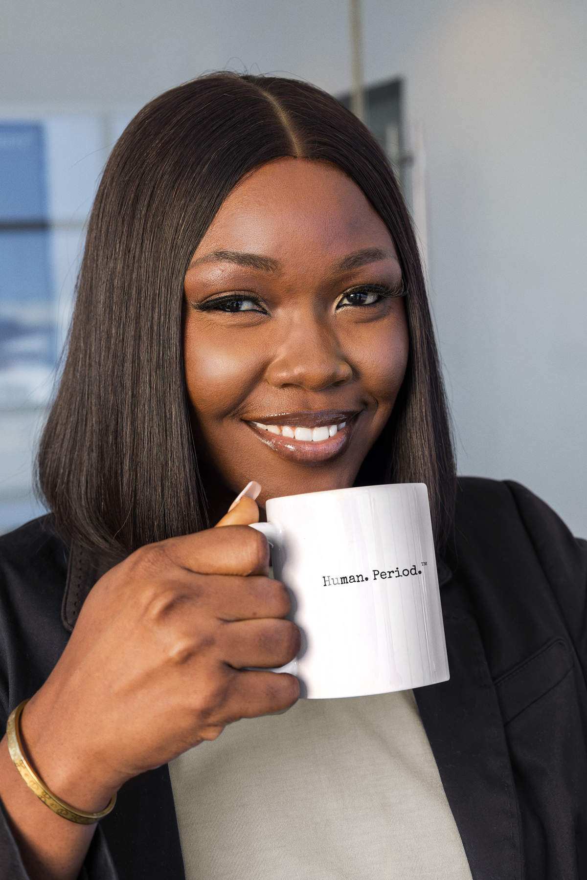 A smiling woman with straight dark hair holds a white ceramic mug printed with the text 'human.Period.