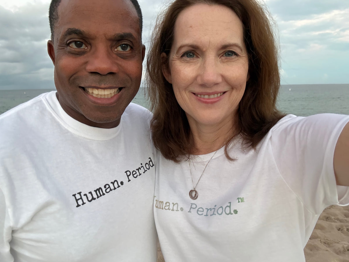 Two people on a beach smiling wearing Human. Period. shirts.