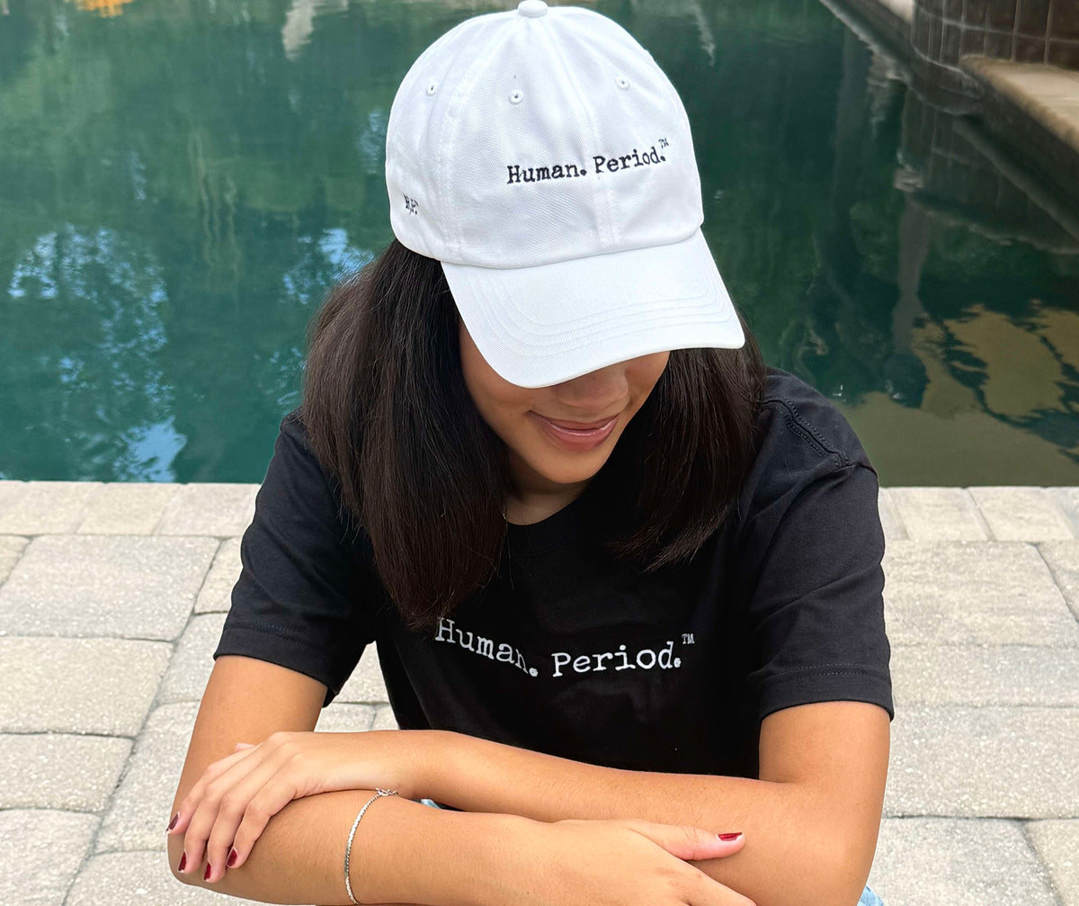 A woman sitting outdoors by a pool wearing a white baseball cap and a black t-shirt, both featuring the text 'Human Period.' in white.
