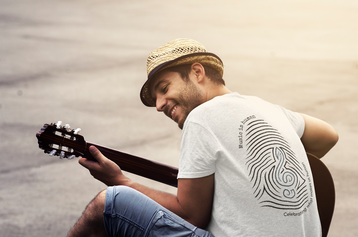 A man wearing a straw hat and a white graphic t-shirt sits and plays an acoustic guitar outdoors against a warm, blurred background.